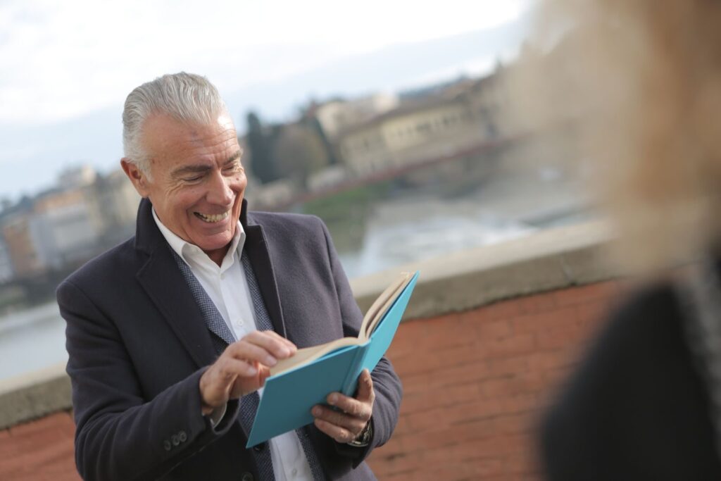 A senior man smiling while reading a book outdoors. Bright and joyful moment.