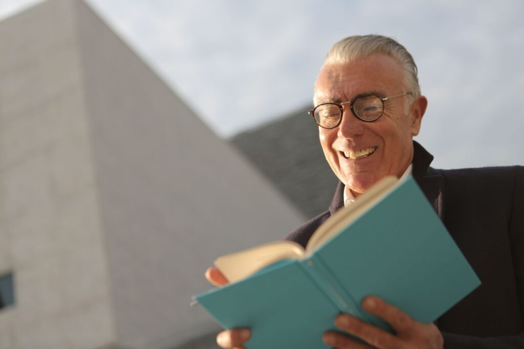 Senior adult reading a book outdoors, smiling under bright daylight.