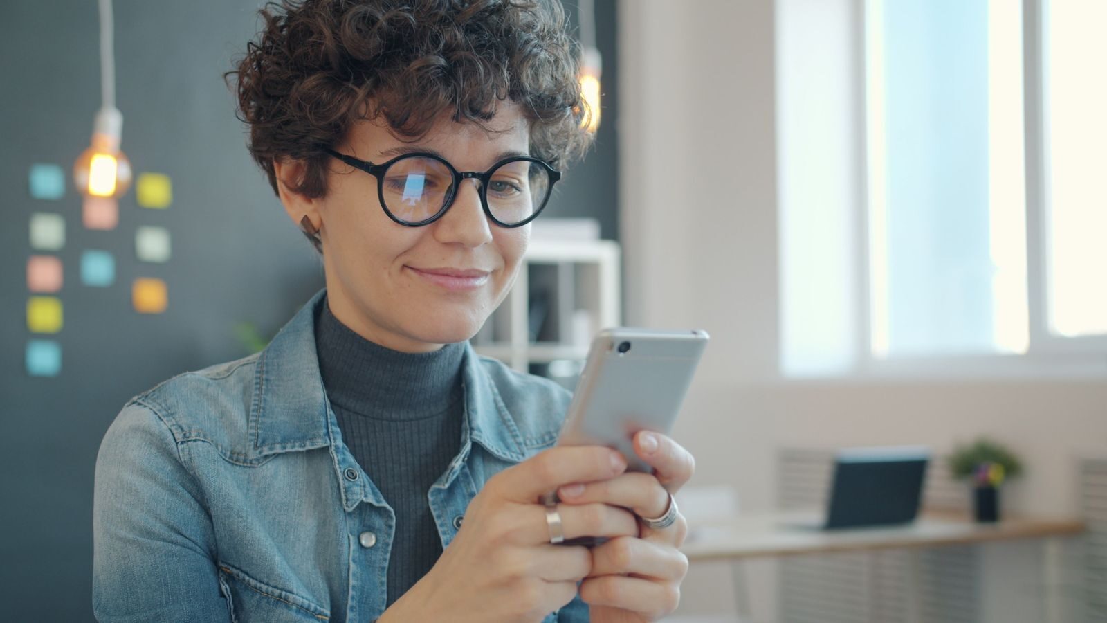 Woman in glasses using smartphone in modern office, smiling at screen.